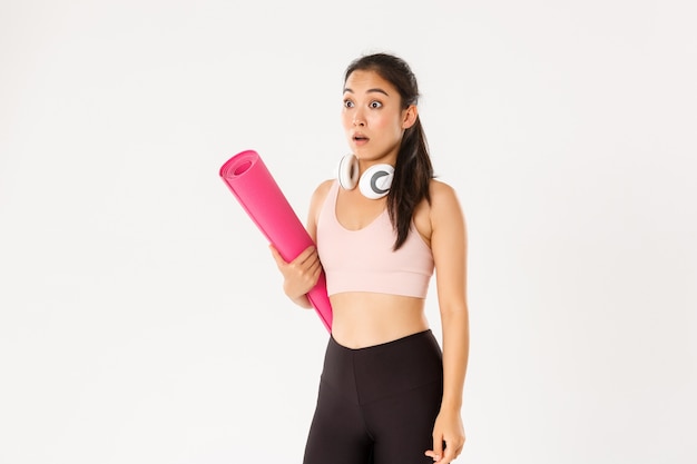 Shocked asian fitness girl in stupor, drop jaw and gasping while looking left at logo or banner info, holding rubber mat for fitness exercises.