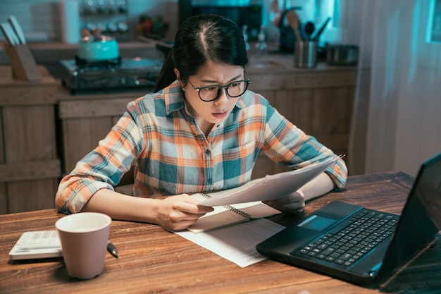 Shocked asian japanese female household paying bills at home
kitchen in dark night. frowning lady surprised by tax document
letter sitting at table with laptop computer. finance problem
concept.