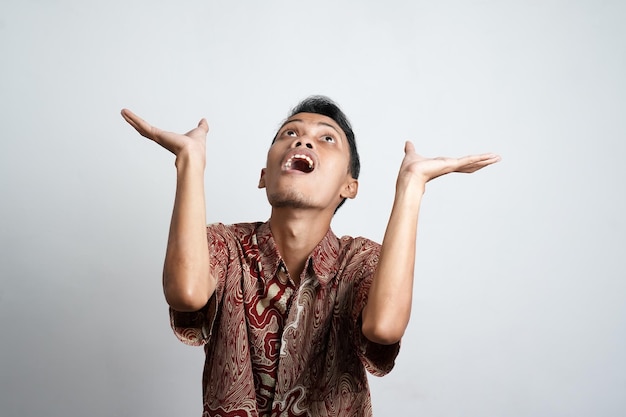 Foto uomo asiatico scioccato che indossa una camicia batik che punta allo spazio della copia sopra di lui isolato da uno sfondo bianco