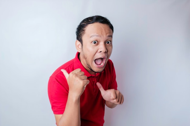 Shocked Asian man wearing a red tshirt pointing at the copy space beside him isolated by a white background