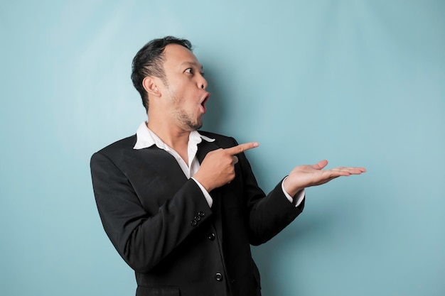 Shocked Asian man wearing suit pointing at the copy space beside him and smiling isolated by blue background