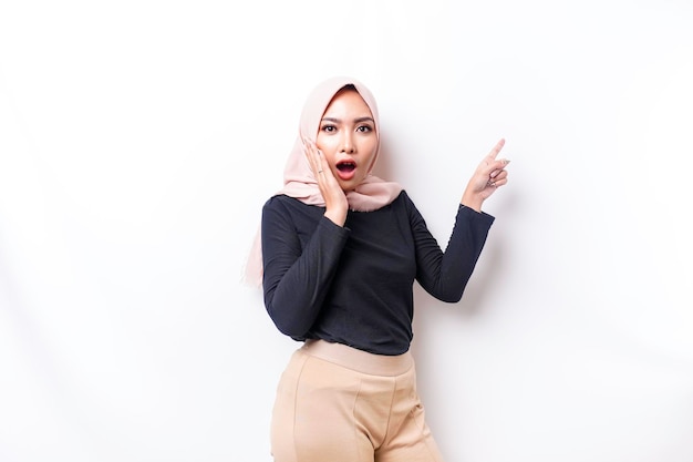 Шокированная азиатская мусульманка, указывающая на пространство для копирования рядом с ней на белом фоне