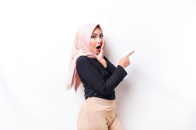 Shocked Asian Muslim woman pointing at the copy space on beside her isolated by white background