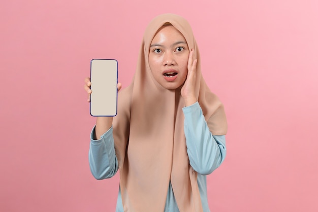 Shocked Asian Muslim Woman touching faces while showing smartphones with blank screen, against on pink background