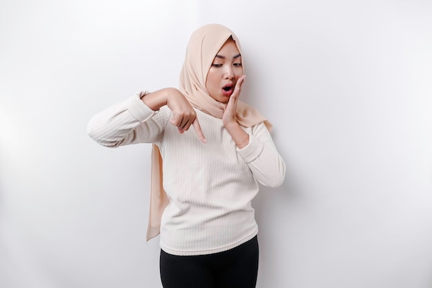 Shocked Asian Muslim woman wearing headscarf pointing at the copy space below her isolated by white background