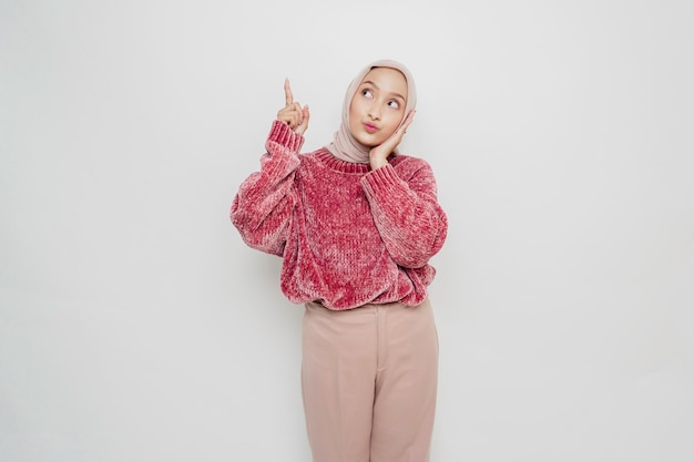 Shocked Asian Muslim woman wearing pink sweater and hijab pointing at the copy space on top of her isolated by white background