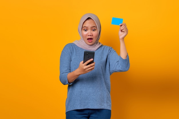 Shocked Asian woman holding mobile phone and showing credit card over yellow background