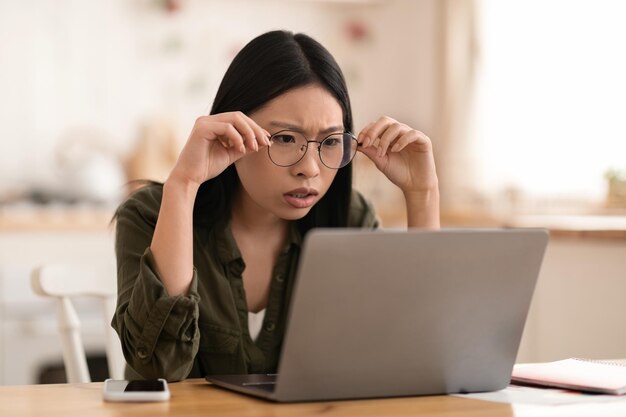 机に座ってノートパソコンを見てショックを受けたアジア人女性