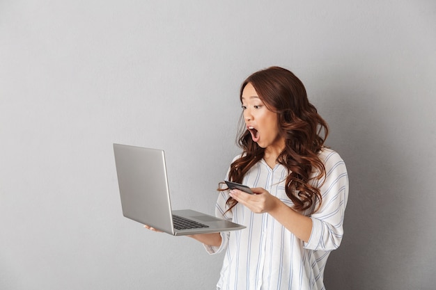 Shocked asian woman standing isolated, looking at laptop computer