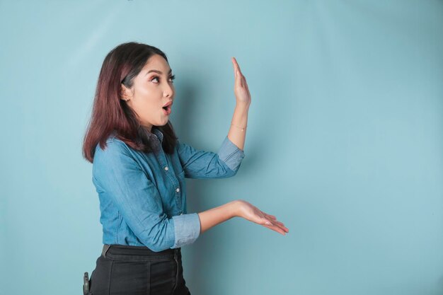 Shocked Asian woman wearing blue shirt pointing at the copy space beside her isolated by blue background