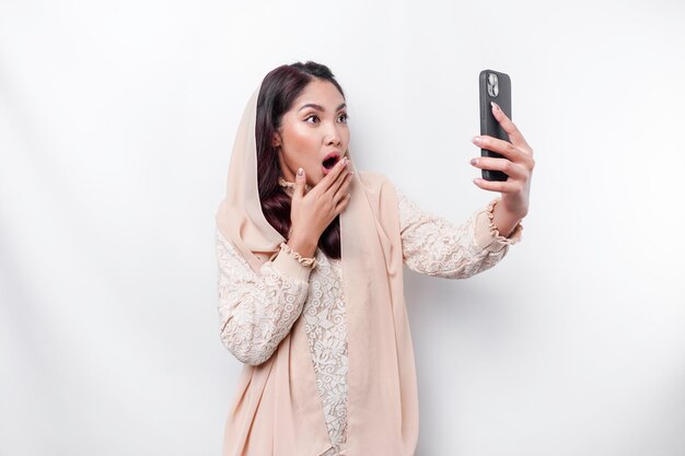 Shocked Asian woman wearing headscarf holding her phone isolated by white background