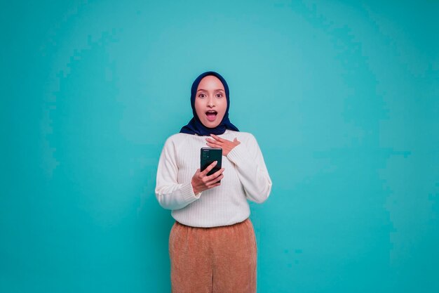 Shocked Asian woman wearing white shirt and hijab holding her phone isolated by blue background