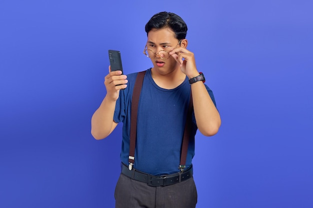 Shocked Asian young man looking at smartphone screen and taking off glasses on purple background