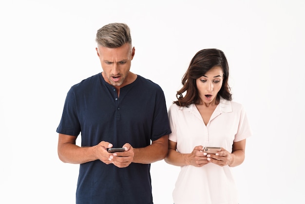Шокированная привлекательная пара в повседневной одежде, стоящая изолированно над белой стеной, используя мобильный телефон