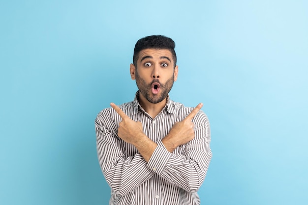 Shocked bearded businessman pointing both sides showing blank
copy space for idea presentation commercial text keeps mouth open
wearing striped shirt indoor studio shot isolated on blue
background