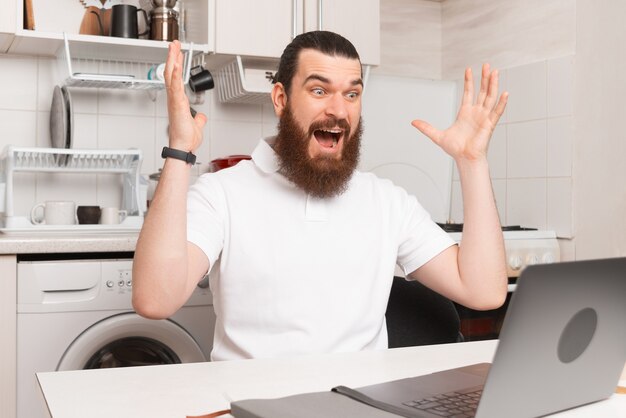 Шокированный бородатый мужчина смотрит на ноутбук, работая за ноутбуком дома.