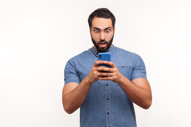 Shocked bearded man looking at smartphone display with big eyes and widely opened mouth extremely surprised amazed with bonuses Indoor studio shot isolated on white background