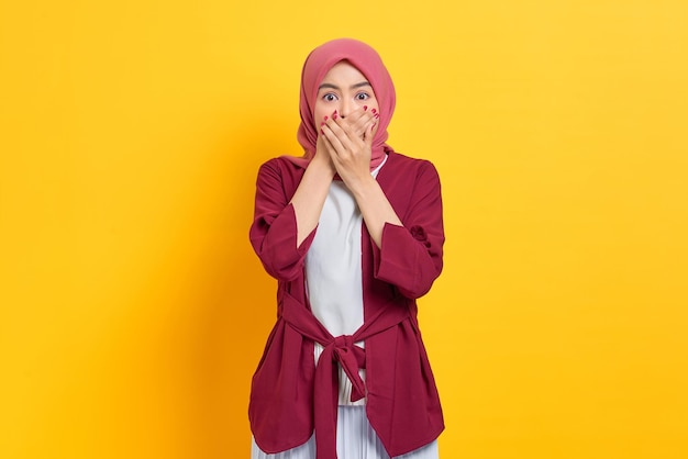 Shocked beautiful Asian woman in casual shirt covering mouth with hands and looking at camera isolated over yellow background