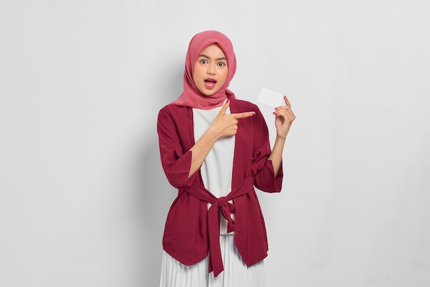 Shocked beautiful asian woman in casual shirt and hijab
pointing fingers at credit card, offer ad product isolated over
white background. people religious lifestyle concept