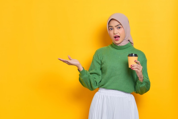 Shocked beautiful Asian woman in green sweater holding paper coffee cup showing copy space on palm isolated over yellow background
