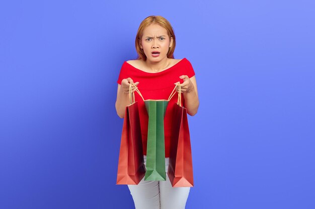 Shocked beautiful Asian woman in red dres opening shopping bags isolated over purple background