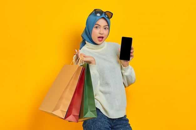 黄色の背景で隔離の空白の画面の携帯電話を示す買い物袋を保持している白いセーターでショックを受けた美しいアジアの女性