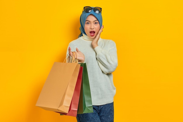 Shocked beautiful Asian woman in white sweater holding shopping bags with open mouth isolated over yellow background