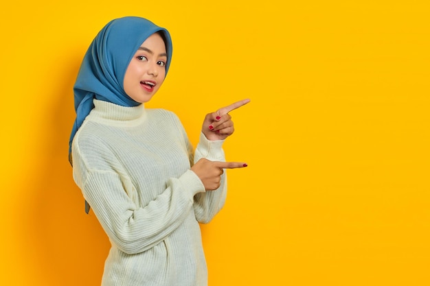 Shocked beautiful Asian woman in white sweater pointing fingers aside at copy space isolated over yellow background