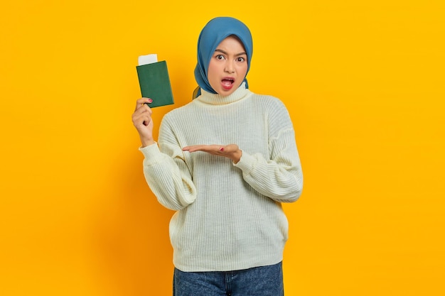 Shocked beautiful asian woman in white sweater showing passport on palm isolated over yellow background summer trip