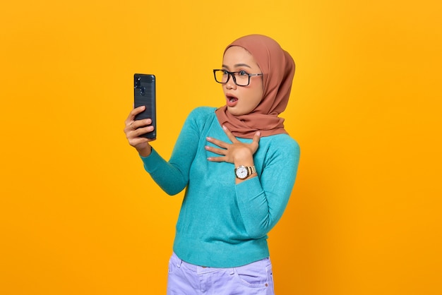 ショックを受けた美しい若いアジアの女性が黄色の背景で隔離のスマートフォン画面を見て