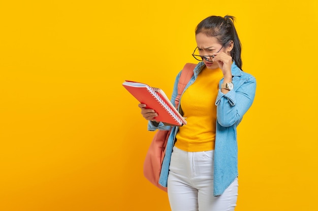 Shocked beautiful young asian woman student in denim clothes\
with backpack reading notebook and looking at task list isolated on\
yellow background education in high school university college\
concept