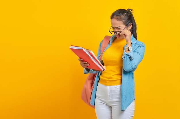 Foto scioccato bella giovane studentessa asiatica in abiti denim con zaino che legge il taccuino e guardando l'elenco delle attività isolato su sfondo giallo istruzione nel concetto di college universitario di scuola superiore