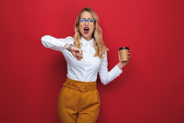 Фото Шокированная красивая молодая блондинка женщина, стоящая изолированно на красном фоне, держа чашку кофе на вынос, проверяя время