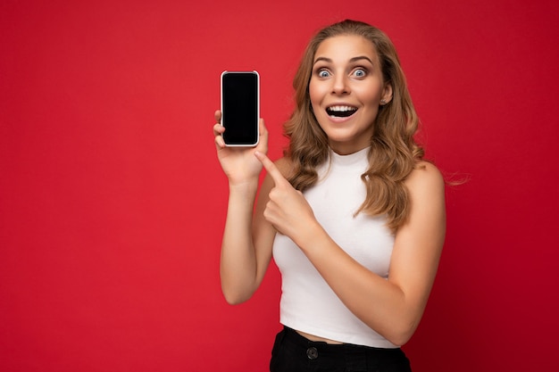 Shocked beautiful young blonde woman with open mouth wearing white tshirt isolated on red background