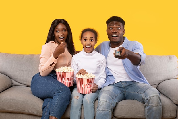 Shocked black parents and their daughter watching movie on TV sitting on sofa over yellow background