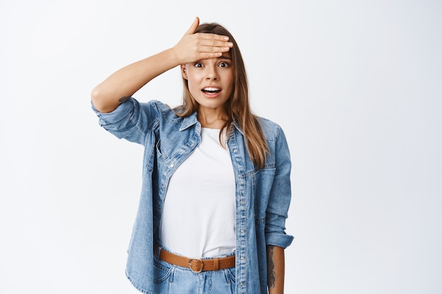 Shocked blond girl slap forehead and staring at front, remember or forget something bad, looking worried, standing in casual clothes against white wall