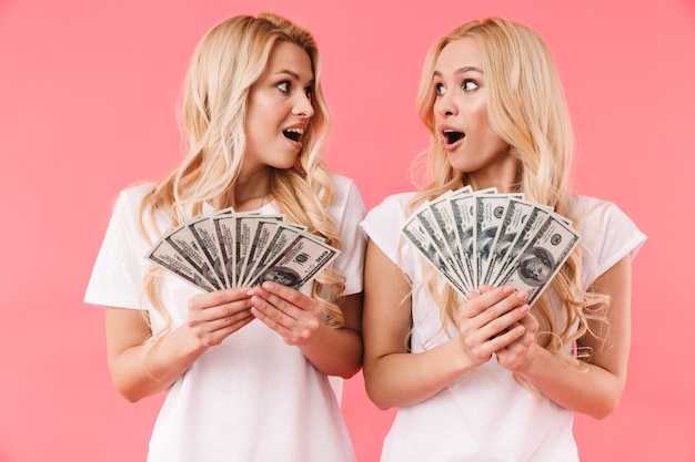 Шокированные блондинки-близнецы в футболках с деньгами, глядя друг на друга на розовой стене