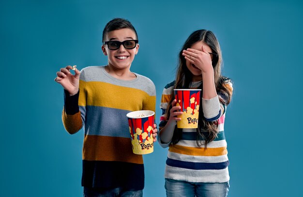 Shocked boy and girl eat popcorn in 3d glasses watching a scary movie isolated on a blue background