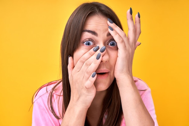 Shocked brunette girl looking surprised with open mouth covers face with hands on yellow background with copy space