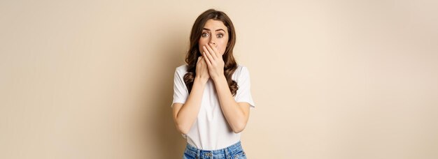 Shocked brunette woman gasping looking speechless at camera cover mouth with hands standing over bei