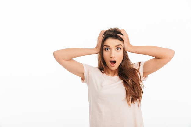 Shocked brunette woman in t-shirt holding her head with open mouth over white wall