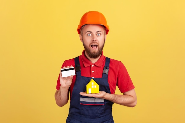 Shocked builder man wearing blue uniform and protective helmet showing credit card and paper house