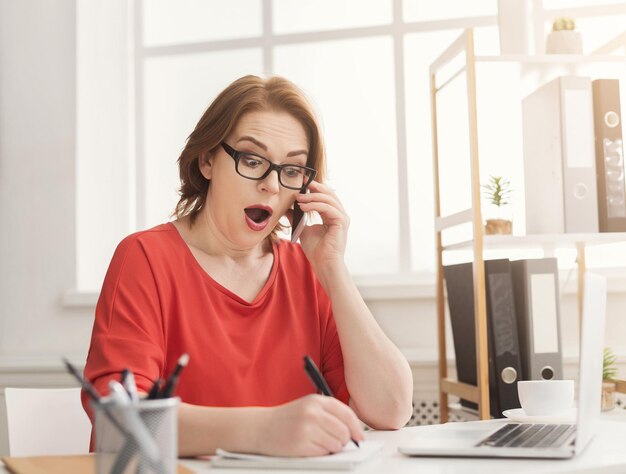 Shocked businesswoman working in office. Financial adviser talking on smartphone and writing to notebook