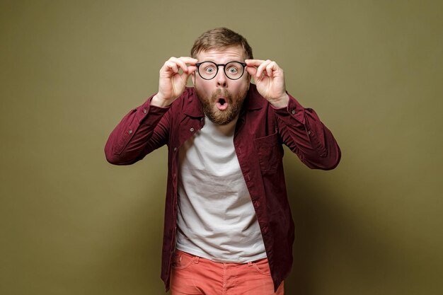 Фото Шокированный кавказский бородатый мужчина в бордовой рубашке держит в руках очки и открывает рот