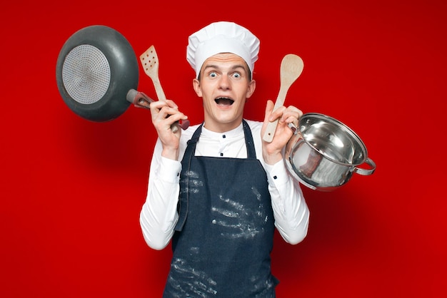 Shocked chef in uniform holds many kitchen items on red isolated background kitchen worker in stress