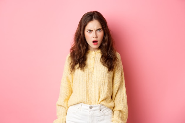 Shocked and confused brunette woman gasping, frowning and looking offended at camera, feeling insulted or displeased, standing over pink wall.