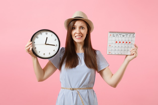 Scioccata confusa donna triste in abito blu con orologio rotondo, calendario dei periodi per controllare i giorni delle mestruazioni isolati su sfondo rosa di tendenza. concetto ginecologico sanitario medico. copia spazio