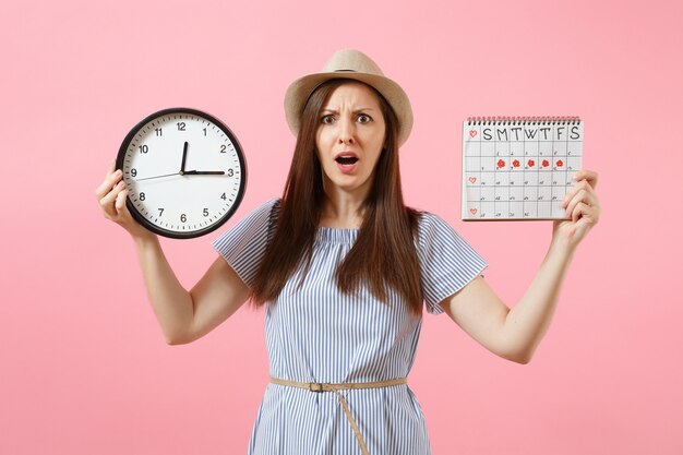 Shocked confused sad woman in blue dress holding round clock, periods calendar for checking menstruation days isolated on trending pink background. Medical healthcare gynecological concept. Copy space