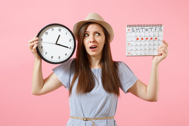 Shocked confused sad woman in blue dress holding round clock, periods calendar for checking menstruation days isolated on trending pink background. Medical healthcare gynecological concept. Copy space