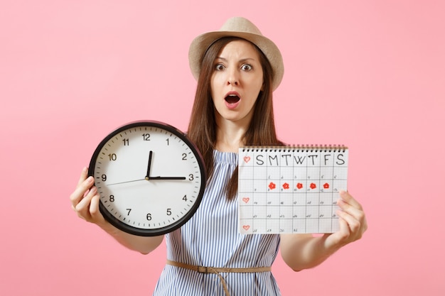 Shocked confused sad woman in blue dress holding round clock, periods calendar for checking menstruation days isolated on trending pink background. Medical healthcare gynecological concept. Copy space